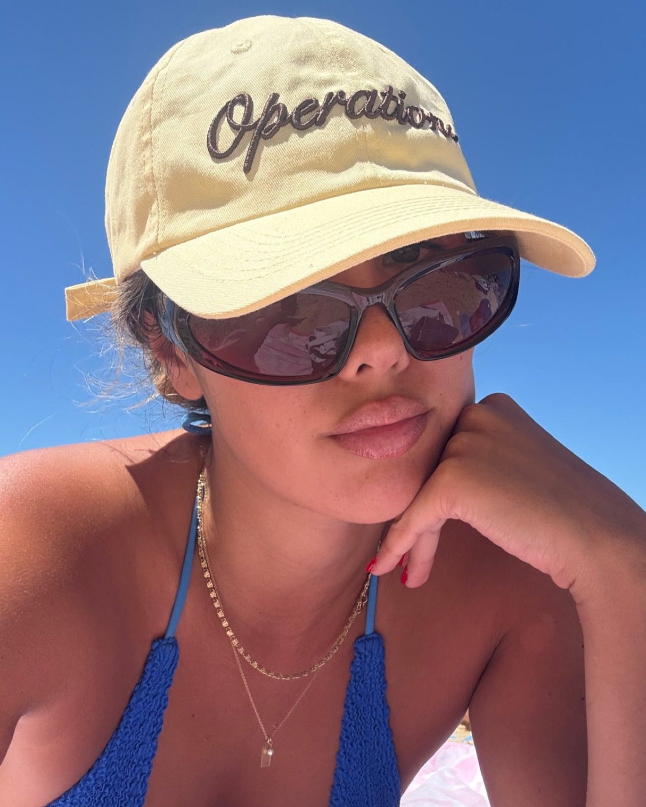 Woman wearing a blue bikini and yellow cap with 'Operation' text, sitting on a beach.